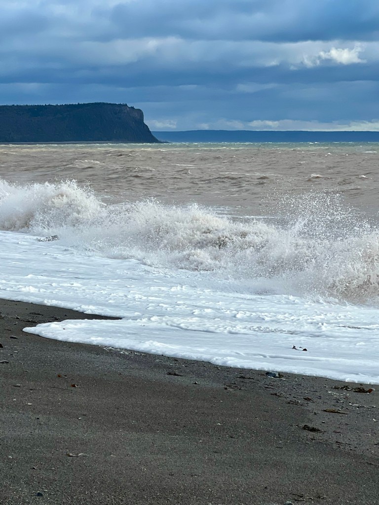 Waves crashing on a sandy beach with a distant cliff and cloudy sky in the background.